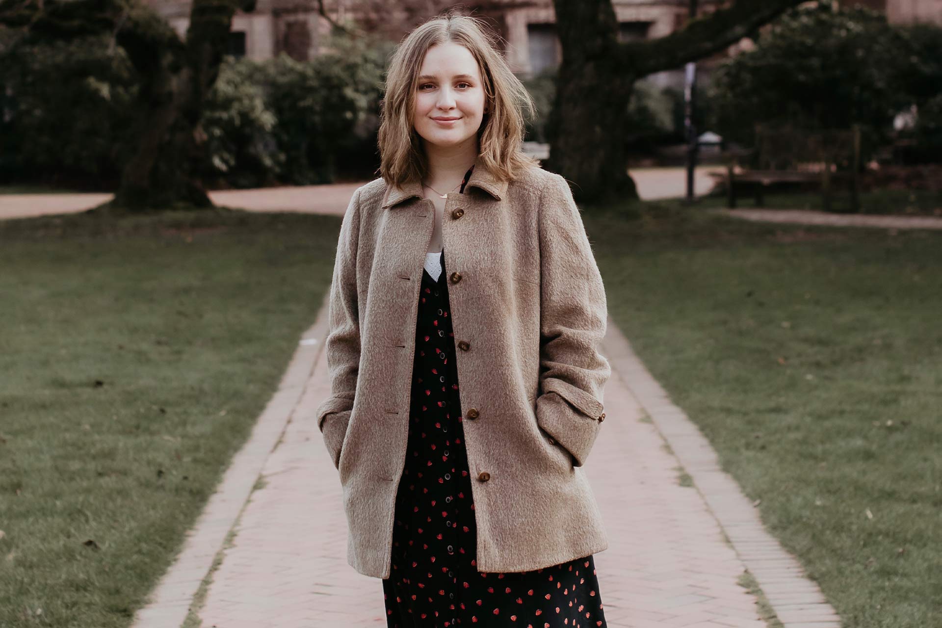 Savannah Miles in a square or park wearing a brown coat smiling at the camera, she has light brown hair.