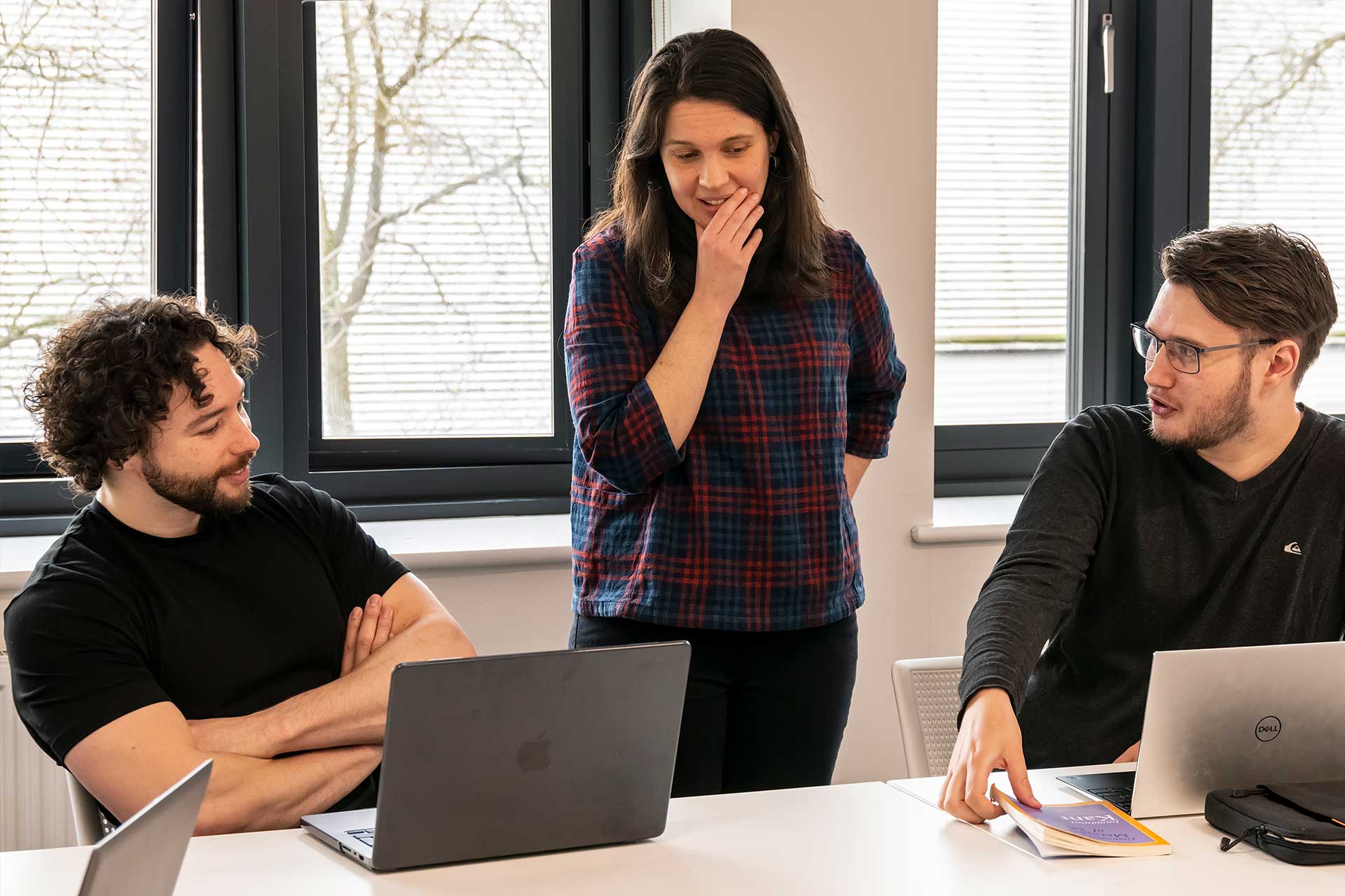 Three people looking at a computer screen. Two men are sat down with laptops and a woman stands in between them looking pensive with her hand on her chin