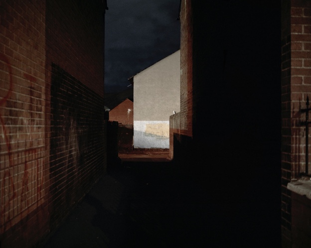 Photorealistic painting or photo of a nighttime alleyway framed by redbrick walls. At the end of the alleyway half of a brighttly illuminated house gable is seen, similar to the way one might see light at the end of a tunnel.