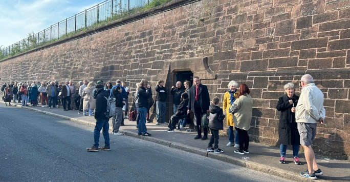 Pohoto of queue of audiecne members alongside the brick wall of Toxteth Reservoir, taken on a sunny day during Beckett Unbound 2024