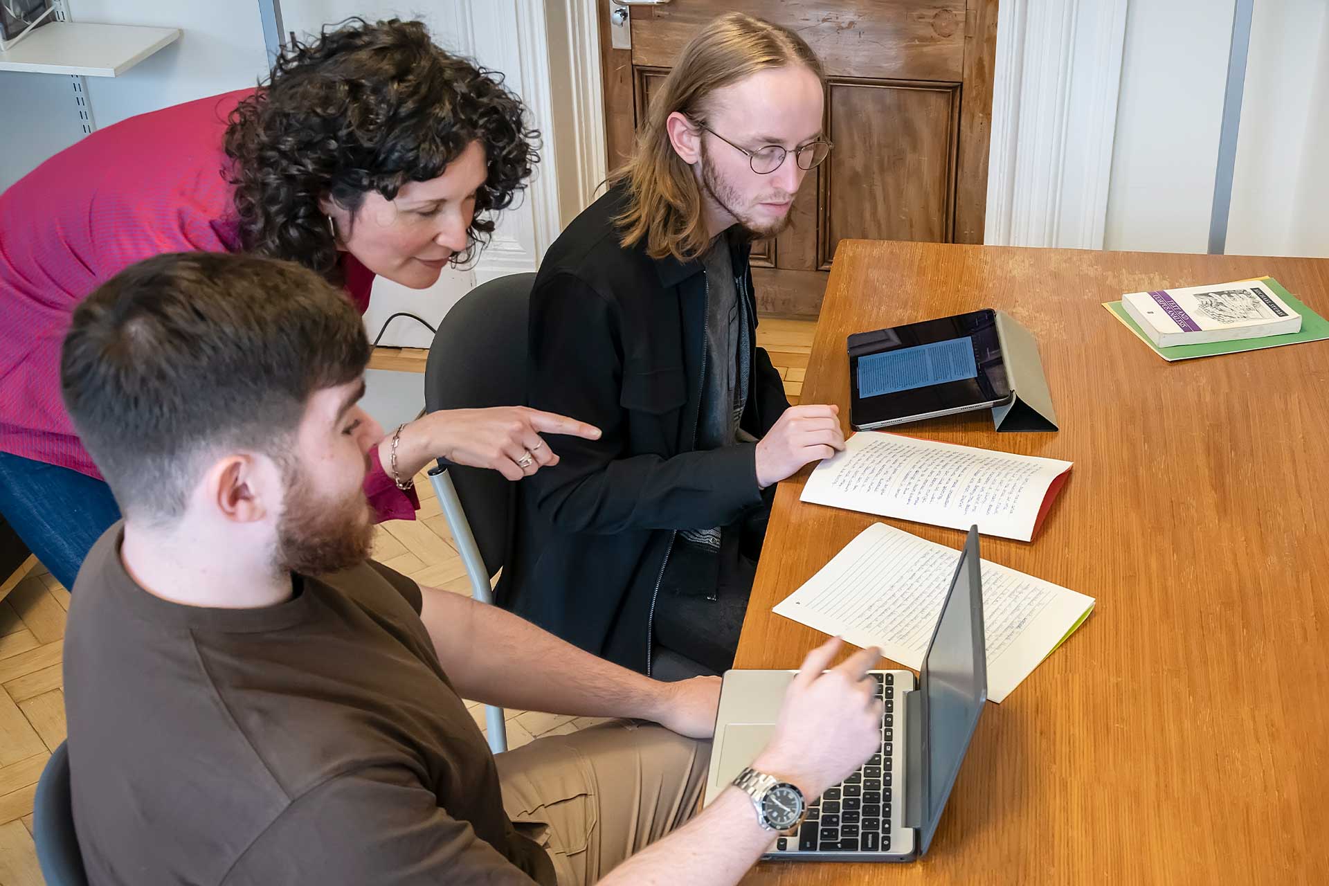 Two students sit at a table with their laptops and a teacher stands behind them pointing at one of their computer screens