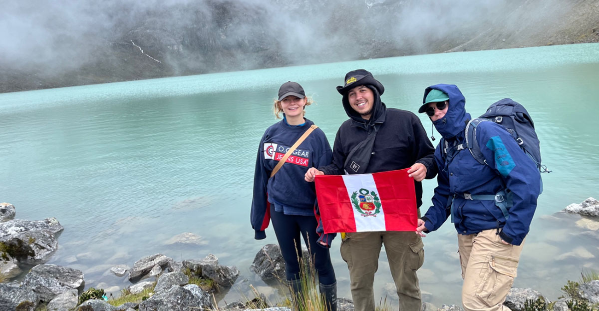 A photograph of three students at a mountain in Peru, they carry the Peruvian flag
