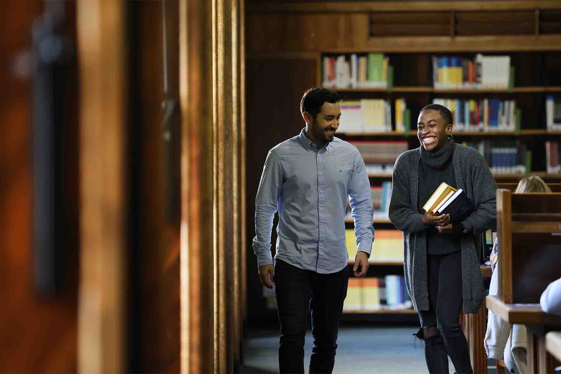 Two students laughing carrying books through the University library.