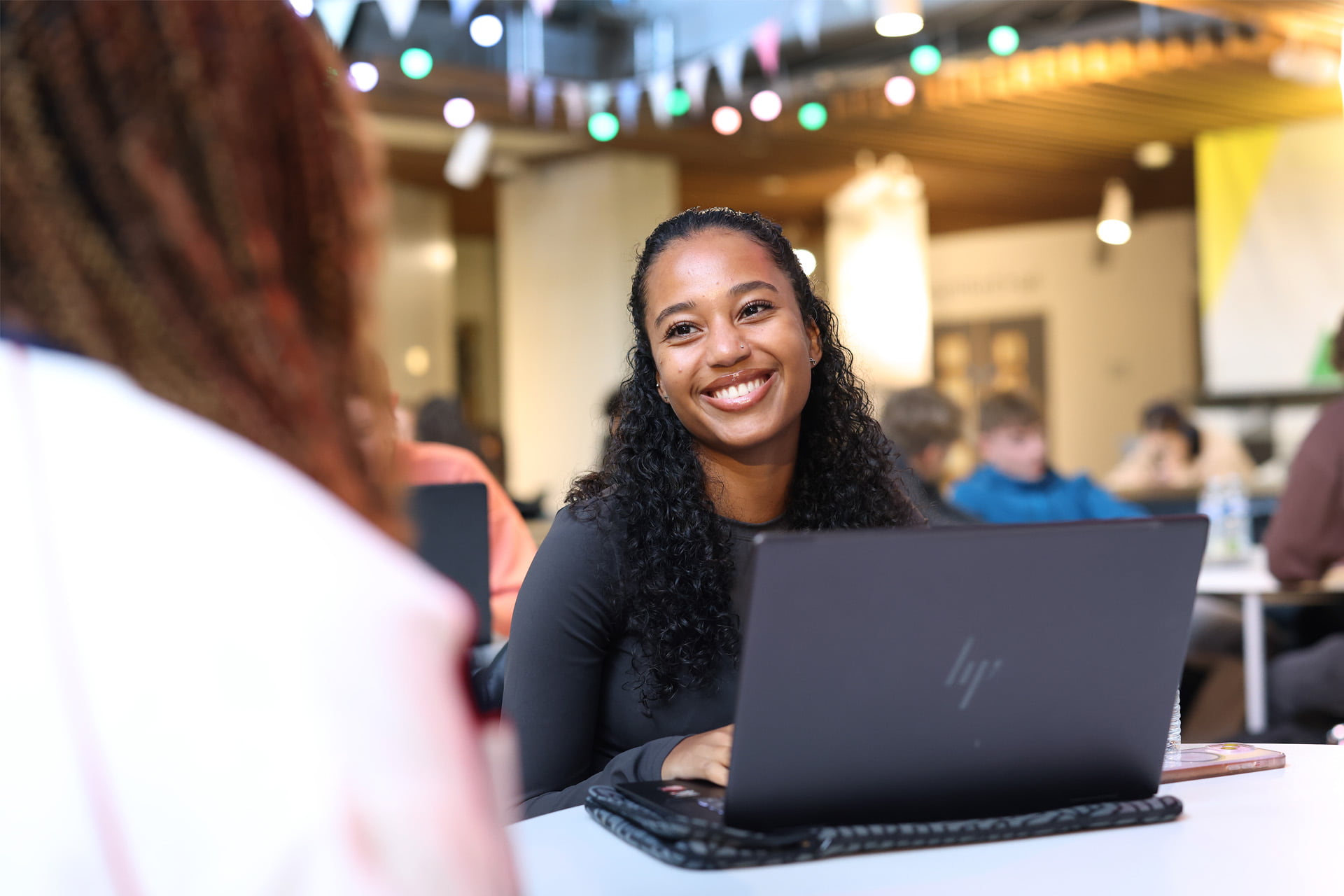 A student sits at a laptop, smiling warmly at someone off-camera.