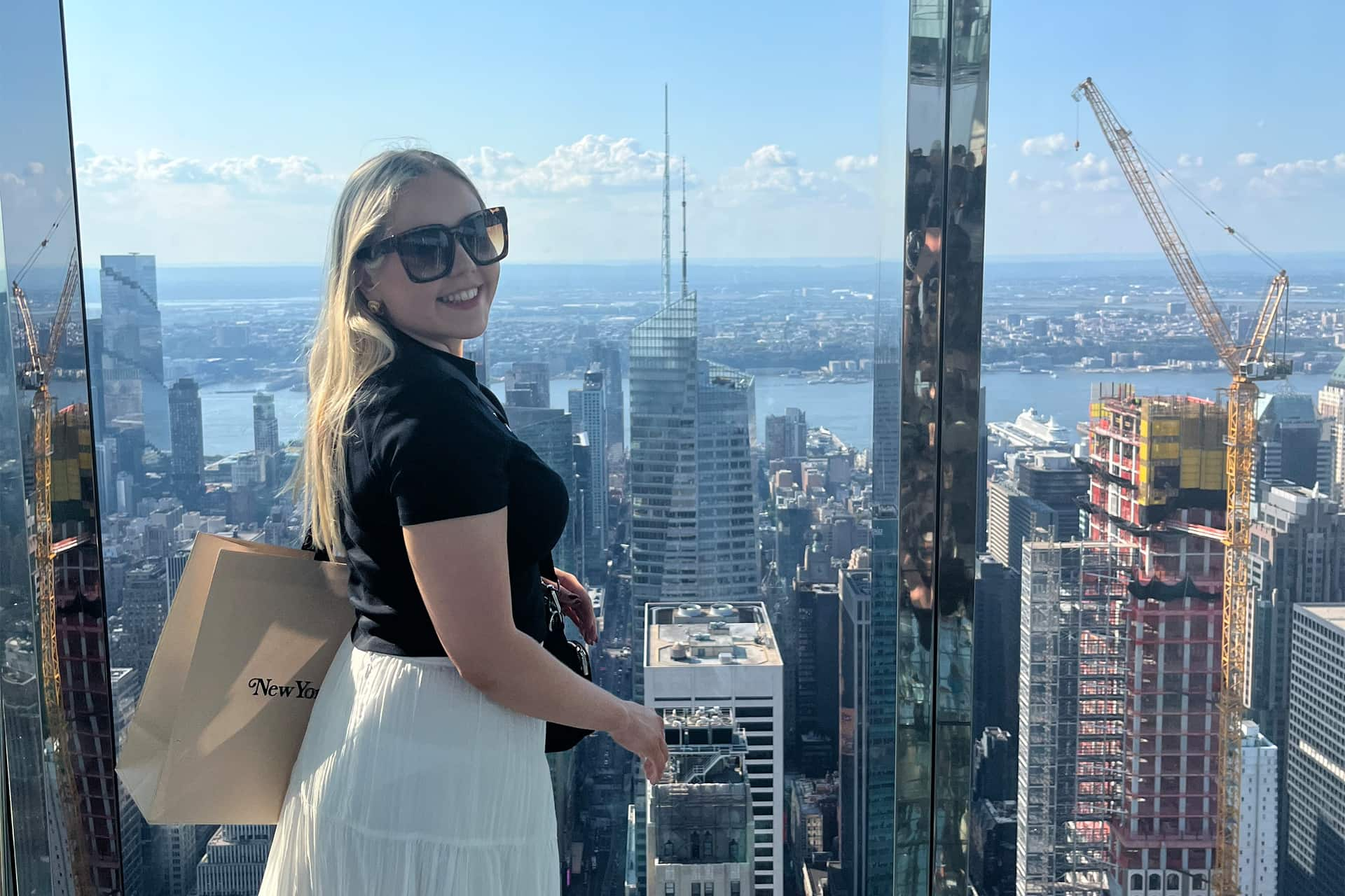 Elizabeth stands at the window of a skyscapper. Behind her is the skyline of Manhattan.