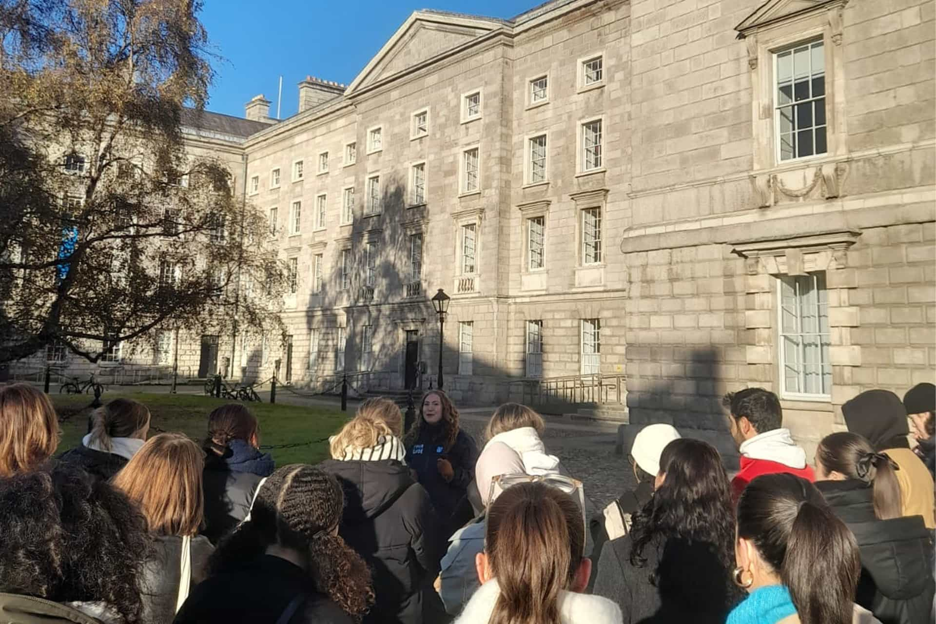 A group of students, led by a tour guide, stand in front of a large grand building.