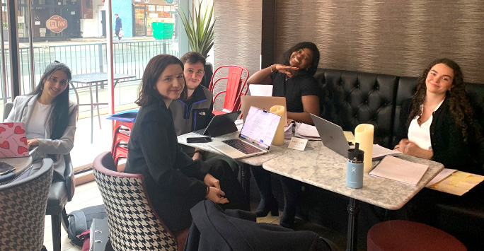 The University of Liverpool Moot Team sit around a table in a cafe together. The table is covered with laptops and paper.