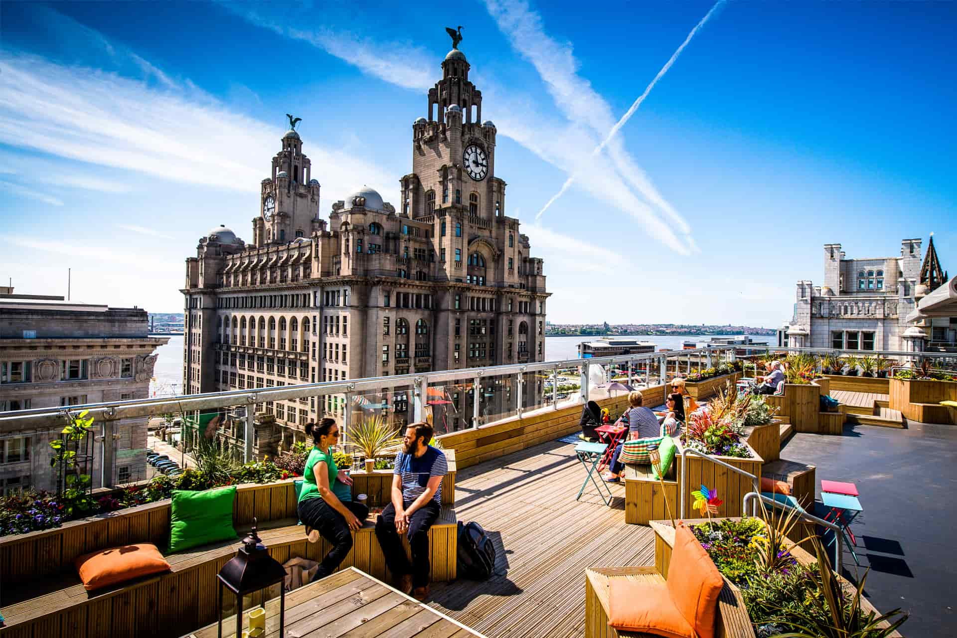 People sitting at a rooftop bar on a sunny day, with the Royal Liver Building in the background.