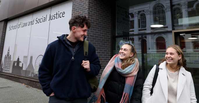 Three students walking together smiling at eachother. The building window it reads 