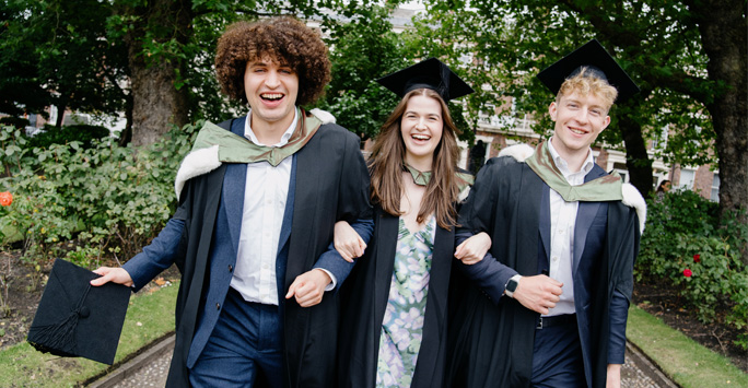 Three students wearing graduation gowns walk together arm in arm.