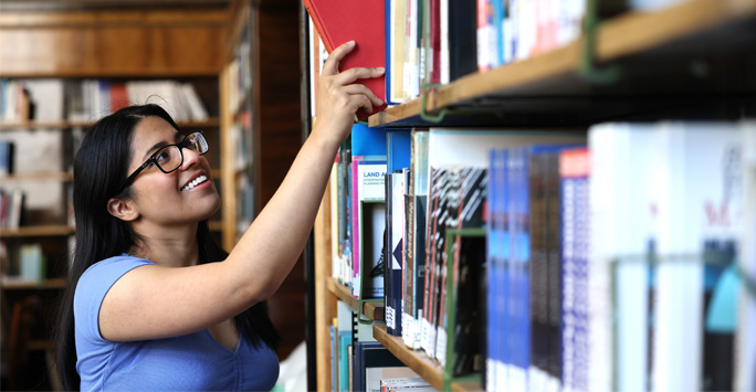 A student reaches for a book from a large bookshelf filled with books.