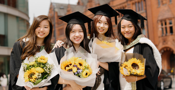 A group of students wearing graduation caps and gowns and holding bunches of flowers, stand with thier arms around each other.