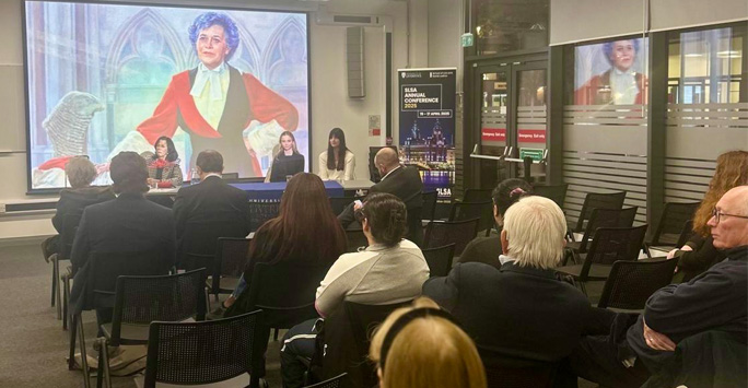 A gathering of individuals in chairs, engaged with a screen presentation which has on it an image of Mrs Justice Heilbron.