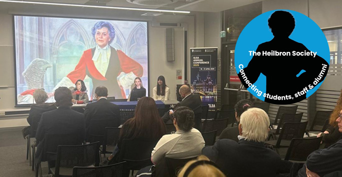 A gathering of individuals in chairs, engaged with a screen presentation which has on it an image of Mrs Justice Heilbron.