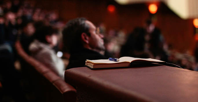 A person listening to a lecture with a notebook in the foreground.