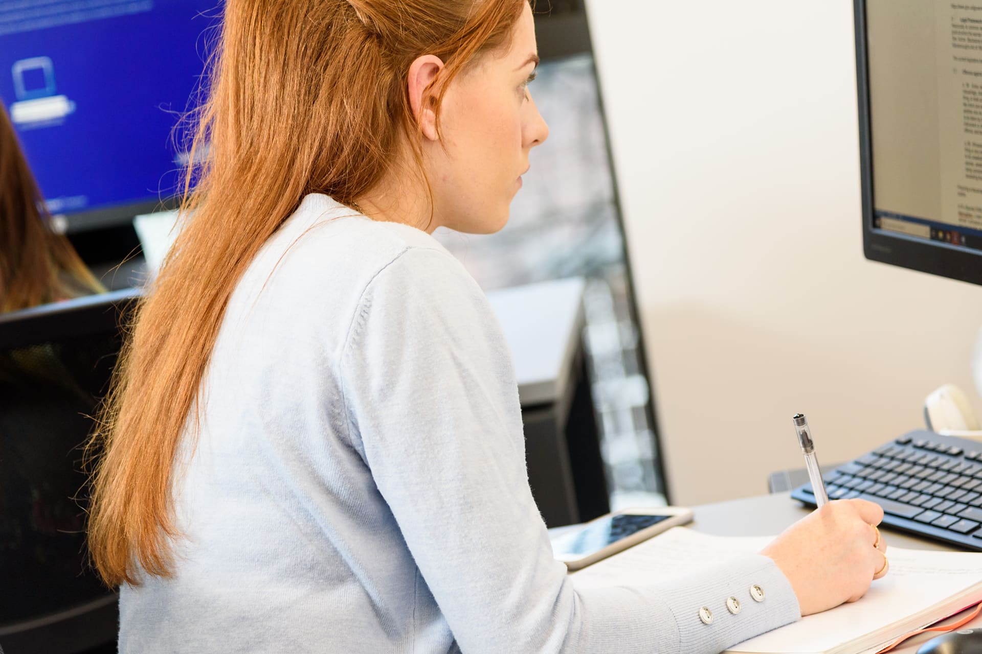 A woman sits at a computer desk making notes.