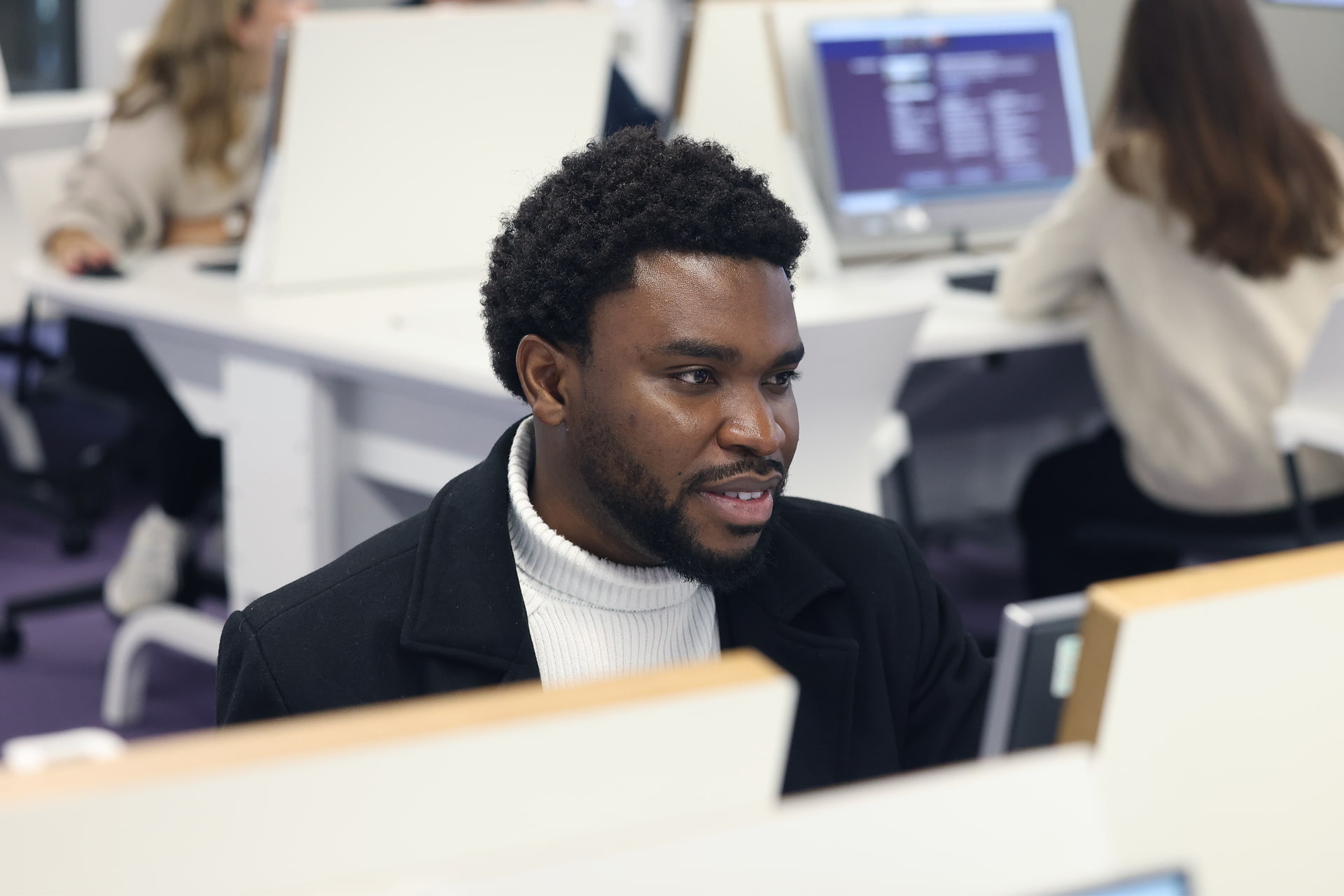 A students sit a computer. In the background there are more computer desks and students.