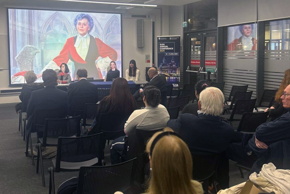 An audience of people sat in chairs. Facing the audience are the panel of speakers. On the screen behind the speakers is a image of Mrs Justice Heilbron.