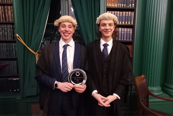 Two students, Danny and Oliver, stand side by side wearing court gowns and wigs, holding a trophy and smiling.