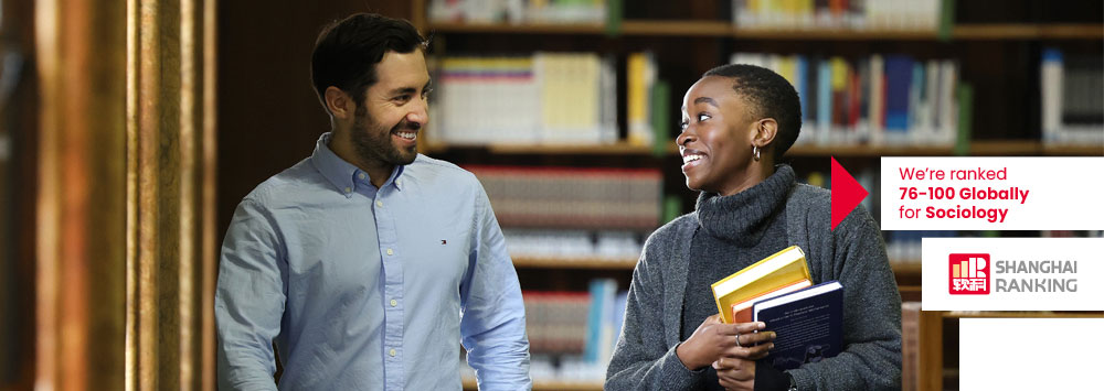 Two students walk through the university library. In the bottom right hand corner is a Shanghai Rankings logo with the text 'We're ranked 76-100 Globally for Sociology'