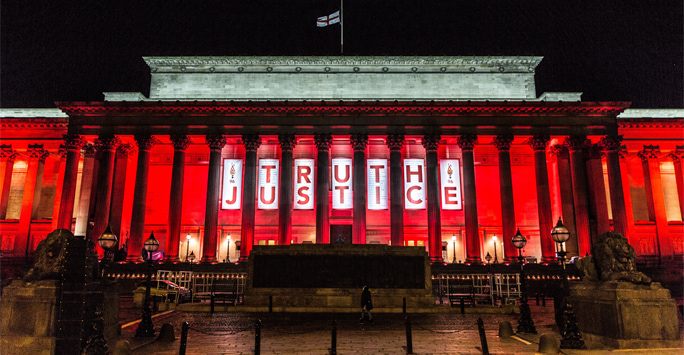 St George's Hall, a grand neoclassical building, illuminated in red at night. A banner at the front displays the words 