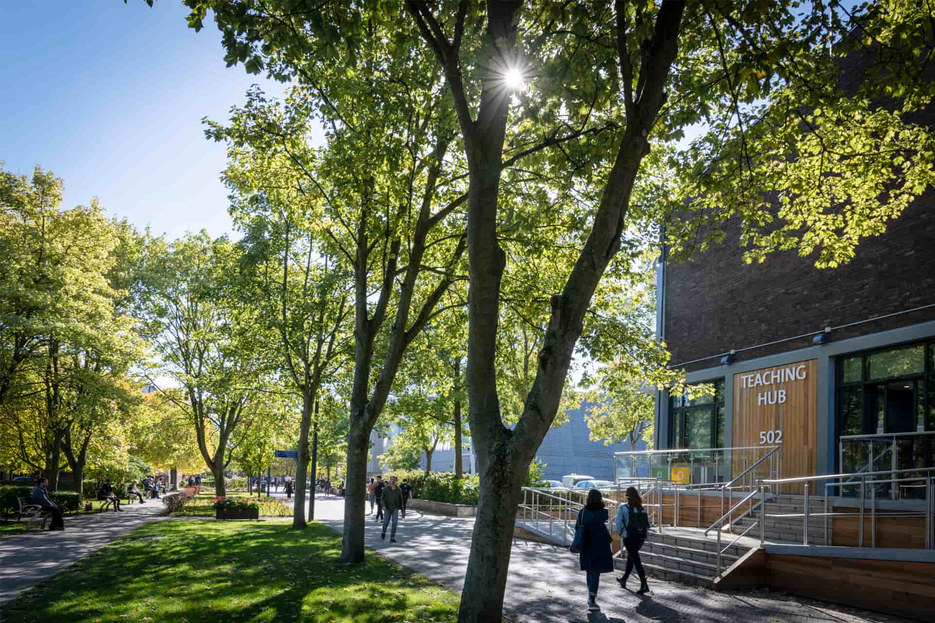 A tree-lined pathway beside buildings, with people strolling along the walkway.