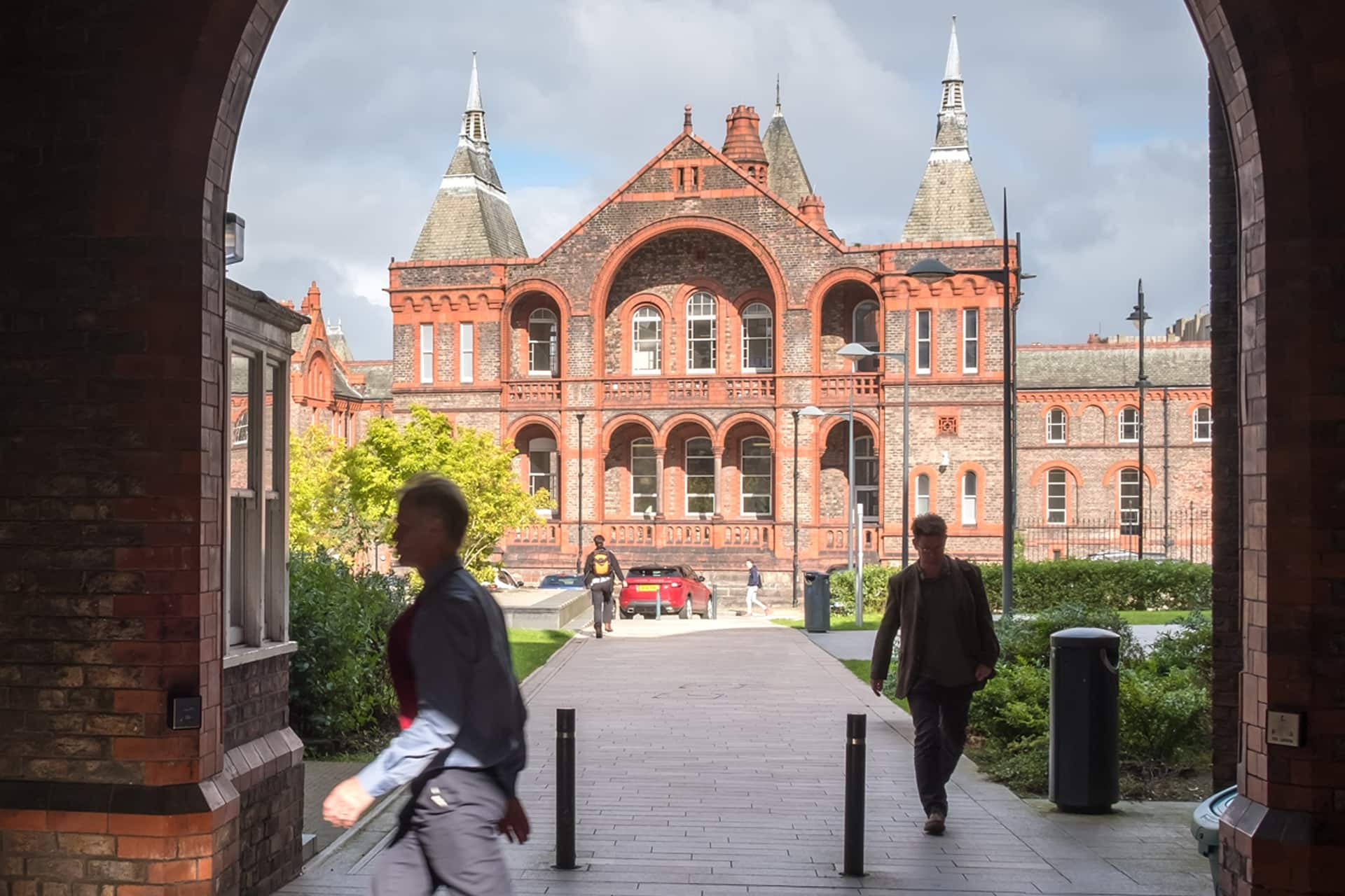 People walking past a large, grand red-brick building with intricate architectural details.