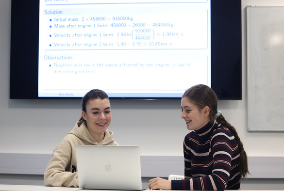 Two students smile while working on a laptop in front of a presentation screen filled with equations and observations.