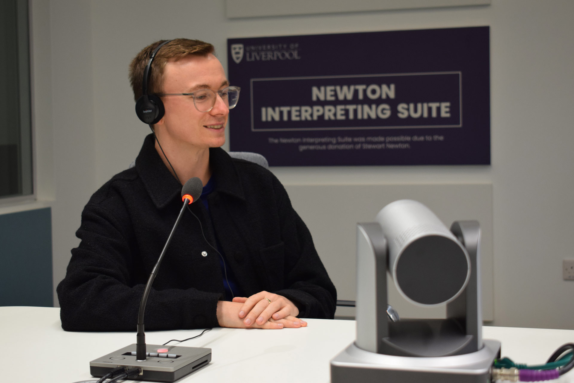 A young man with glasses sits at a desk wearing headphones in front of a microphone, in the background a sign says 'Newton Interpreting Suite'