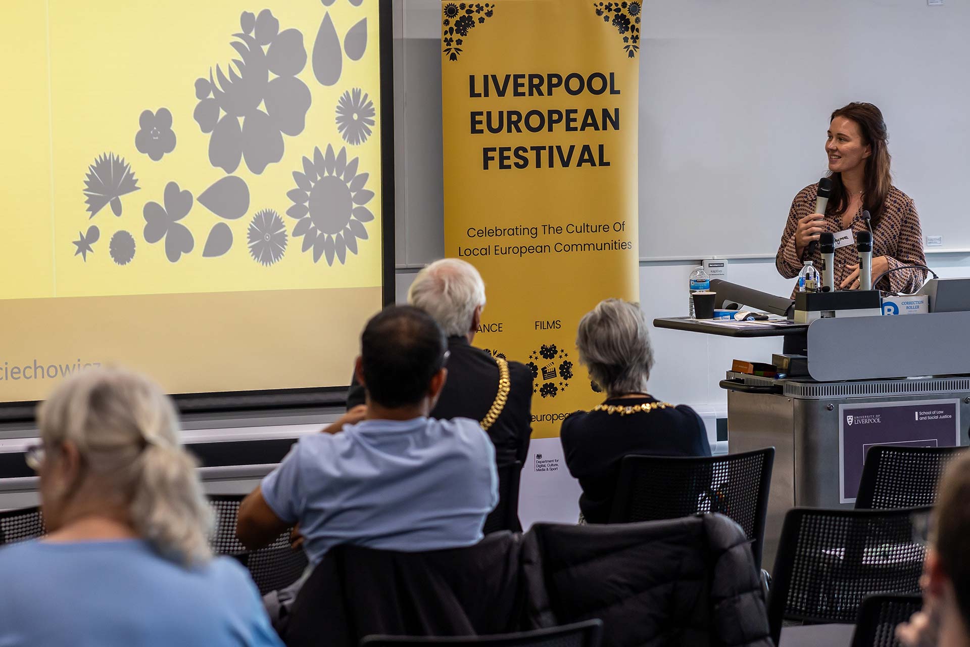 A woman standing at the front of a room with a microphone presenting to a group of people seated.