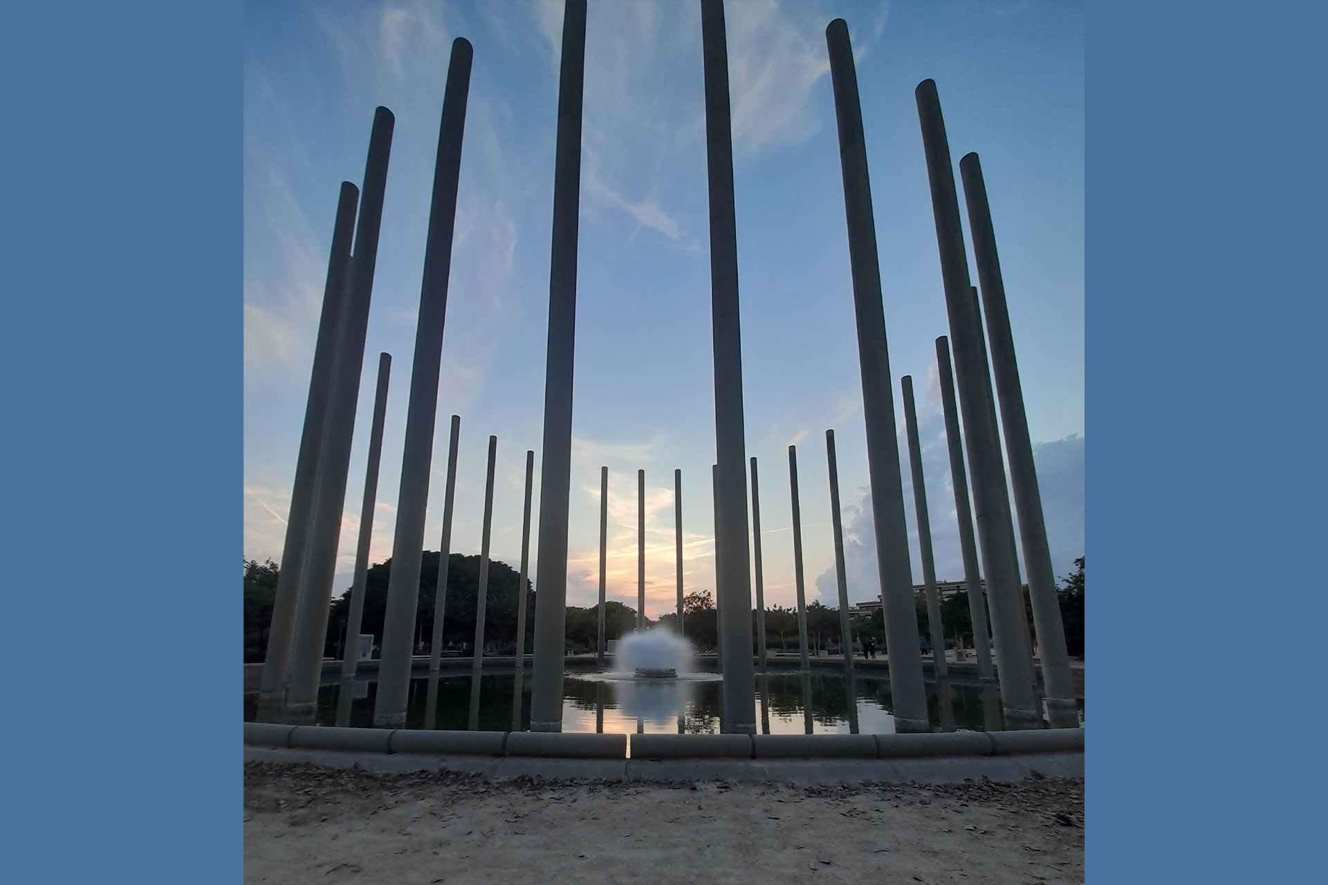 A statue with tall spires arranged in a circle in a park, with a sun setting in the background