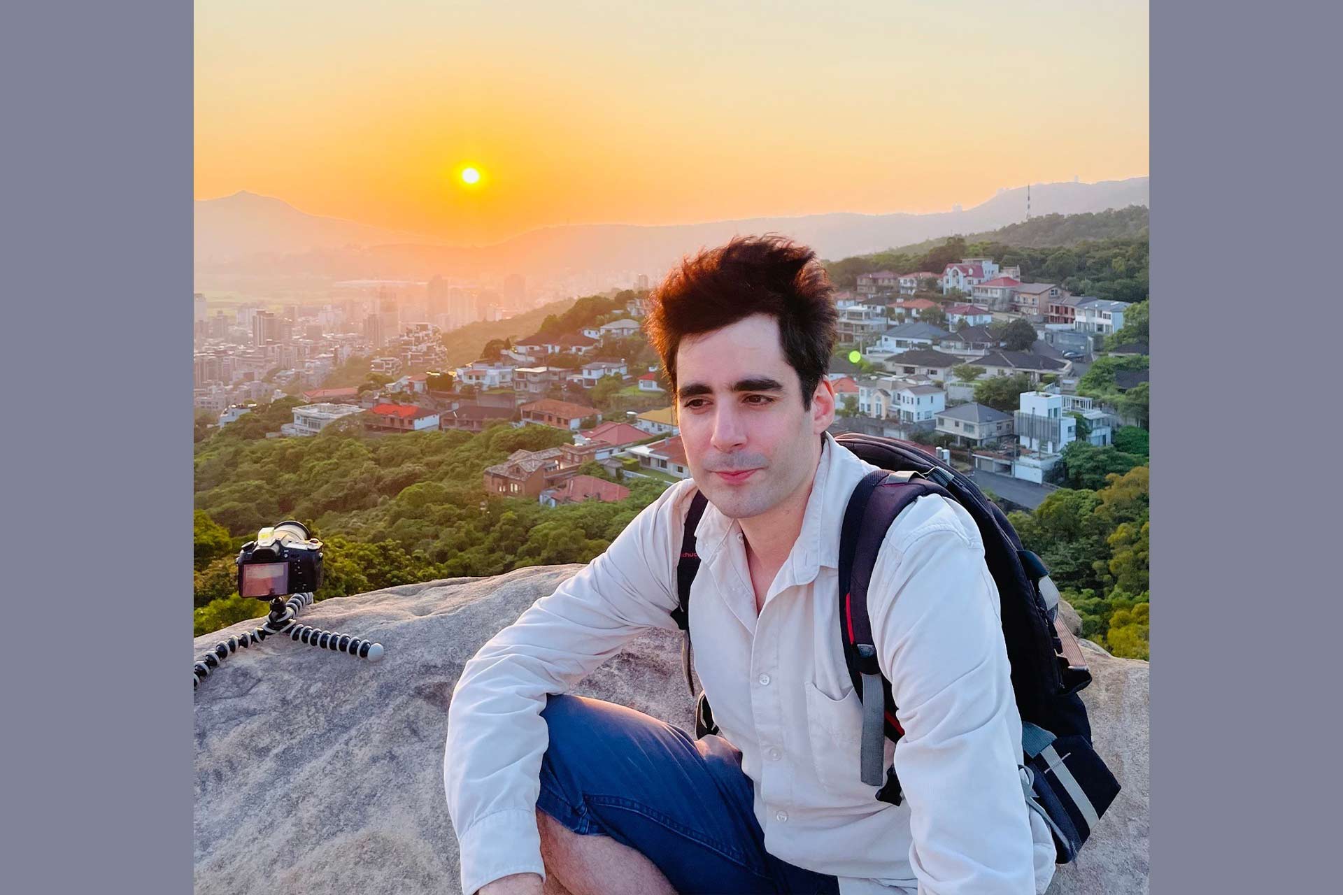 A man sitting on a rock high up above a town, with a sunset in the background