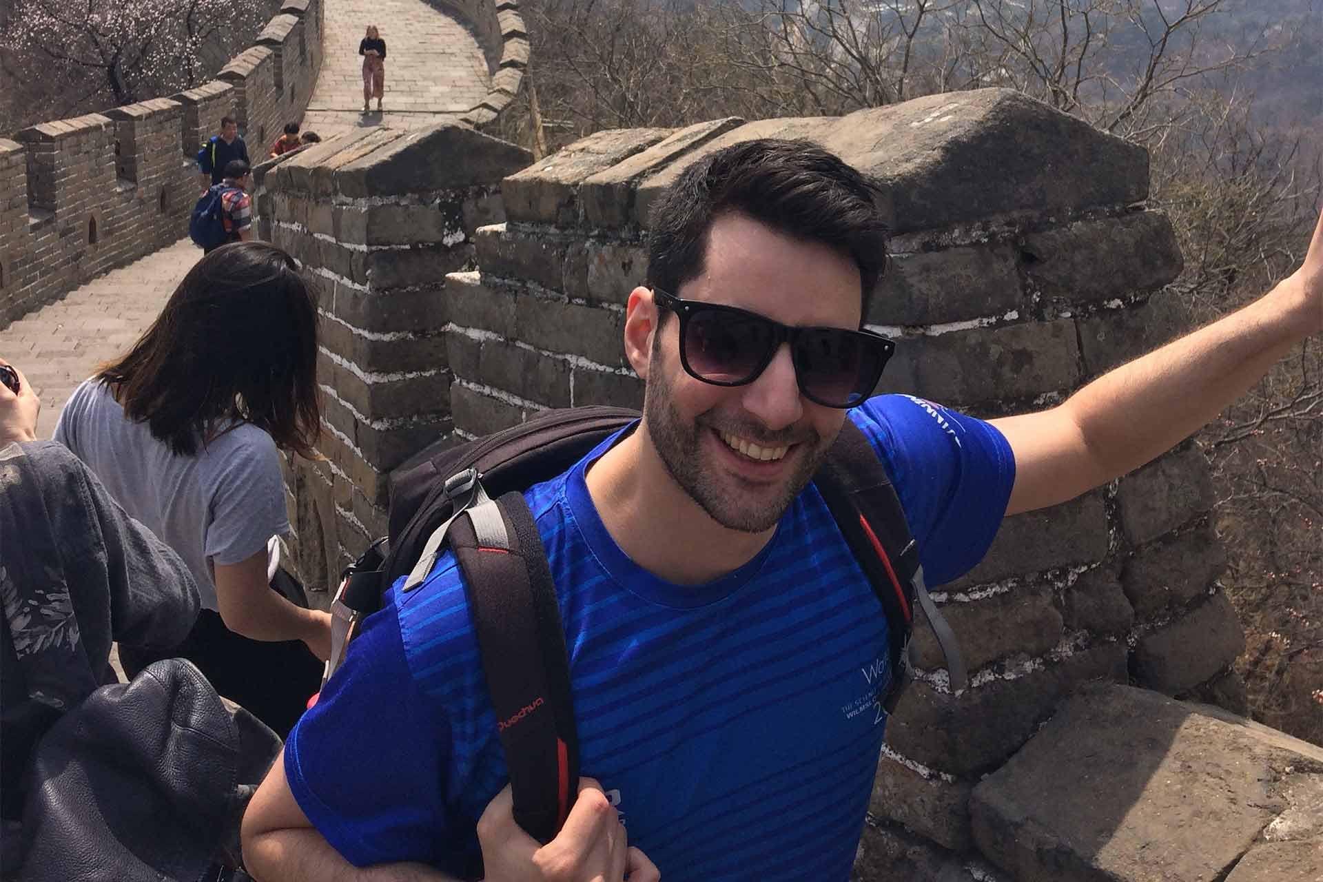 A man wearing a rucksack and sunglasses smiling at the camera standing on the great wall of china