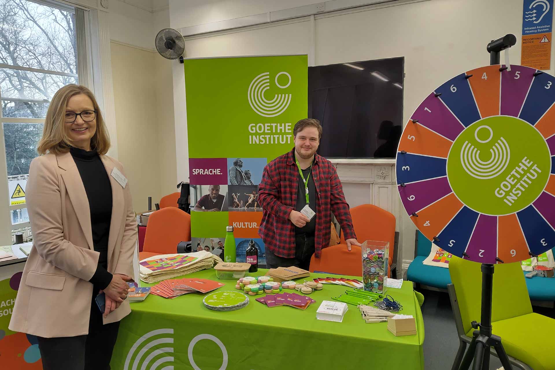 A table is set up with colourful bits of merchandise, a bright green banner in the background reads \'Goethe Institut\'. A woman with blonde hair stands on the left next to the table and younger man with dark hair stands behind the table smiling