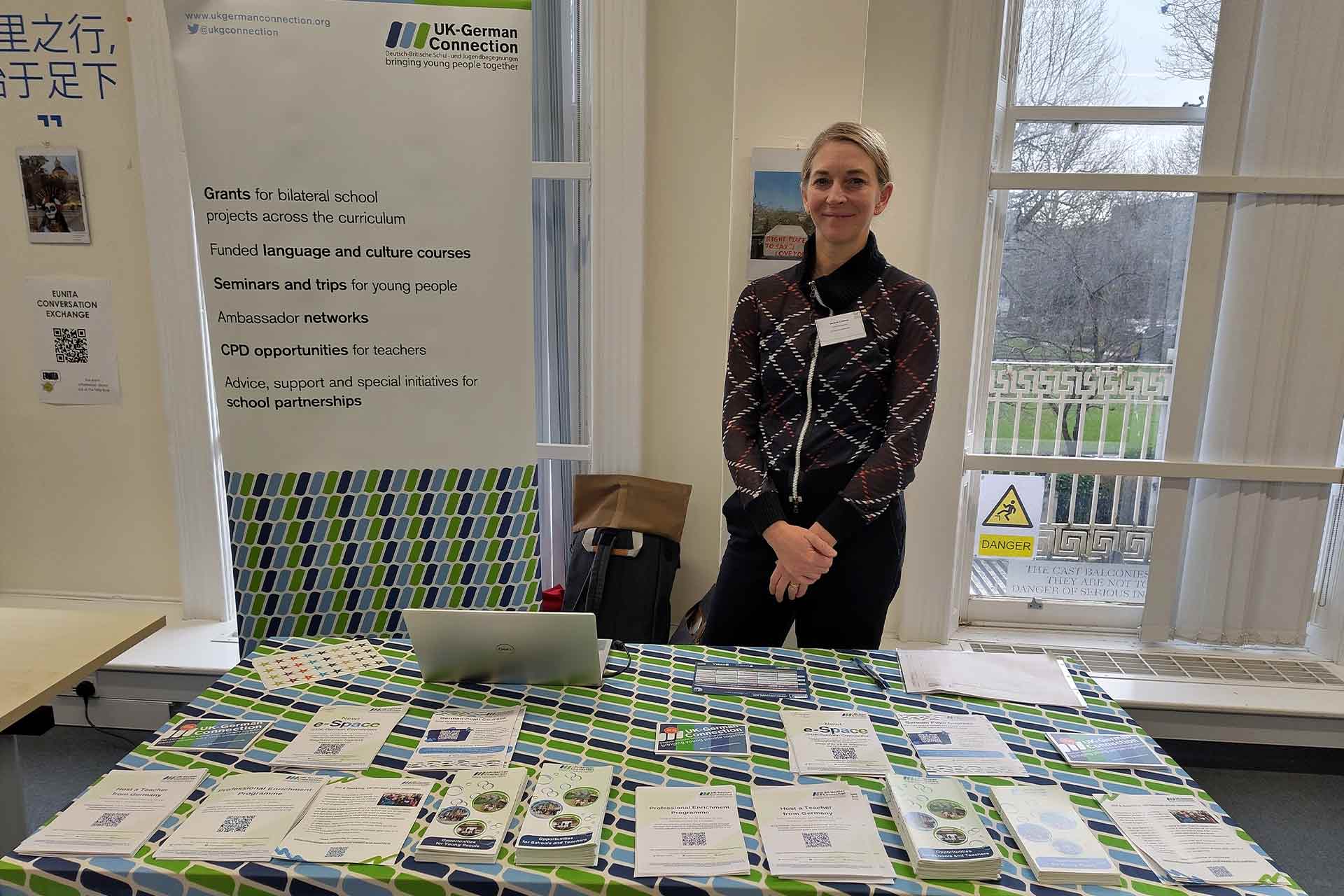 A woman standing and smiling in front of a table with merchandise laid out, a banner behind reads UK-German connection