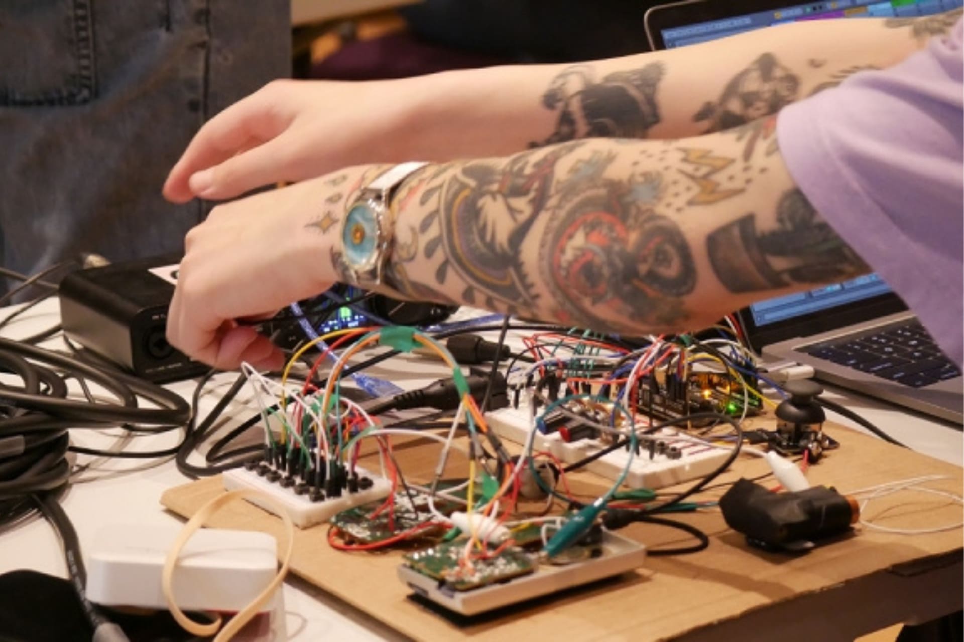 Two tattooed arms are hovering over a circuit board adjusting the colourful wires