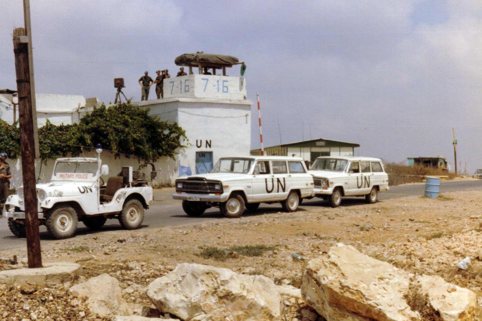 UN personnel stand on top of a building in front of three white UN vehicles in a sandy and rocky terrain.