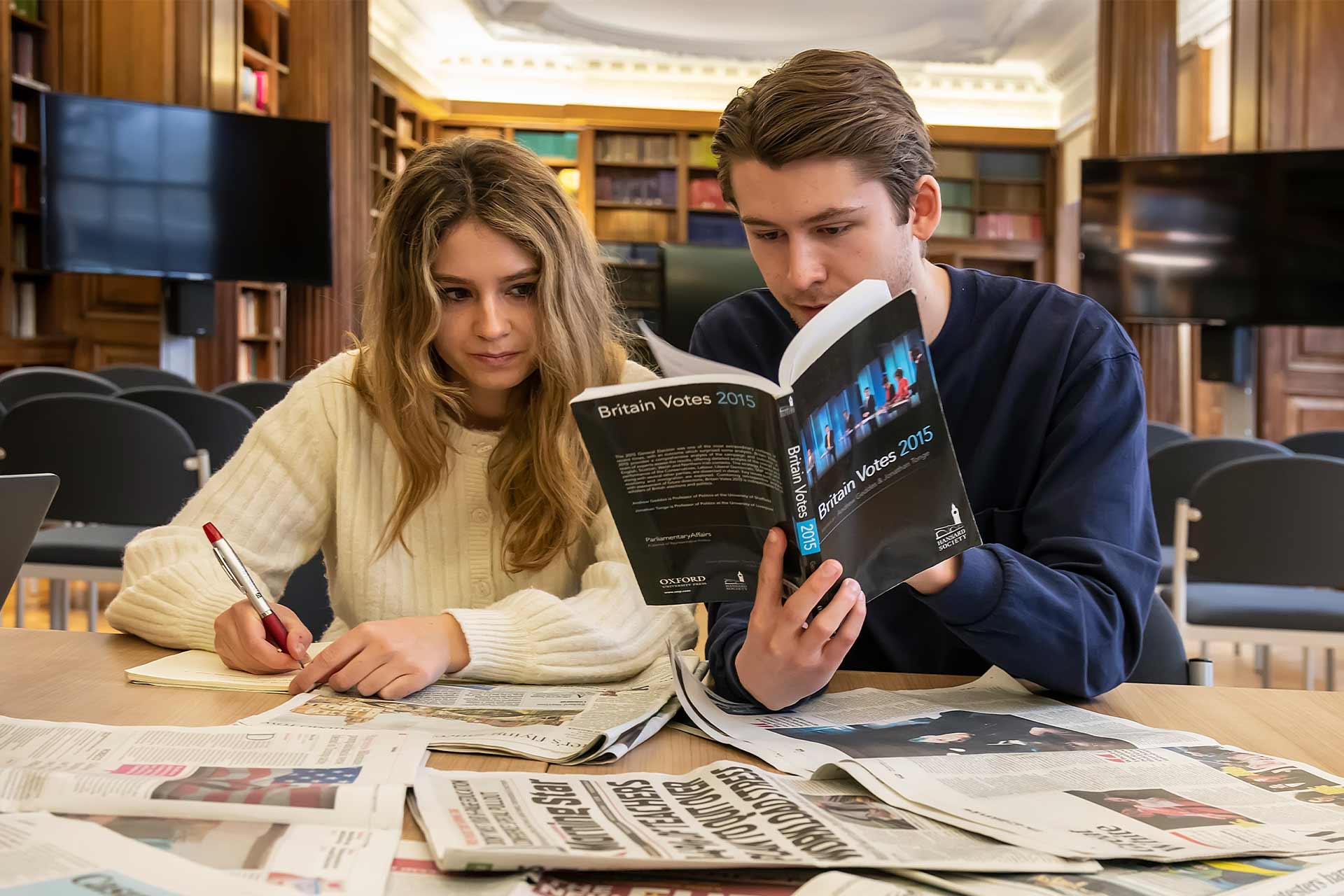 Two students sitting in a library with newspapers spread out on the table in front of them, one student is holding a book entitled 'British Votes 2015'