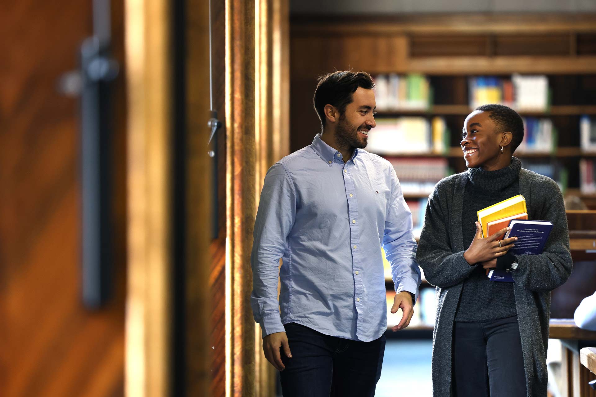 Two students walking through the library holding books and laughing together