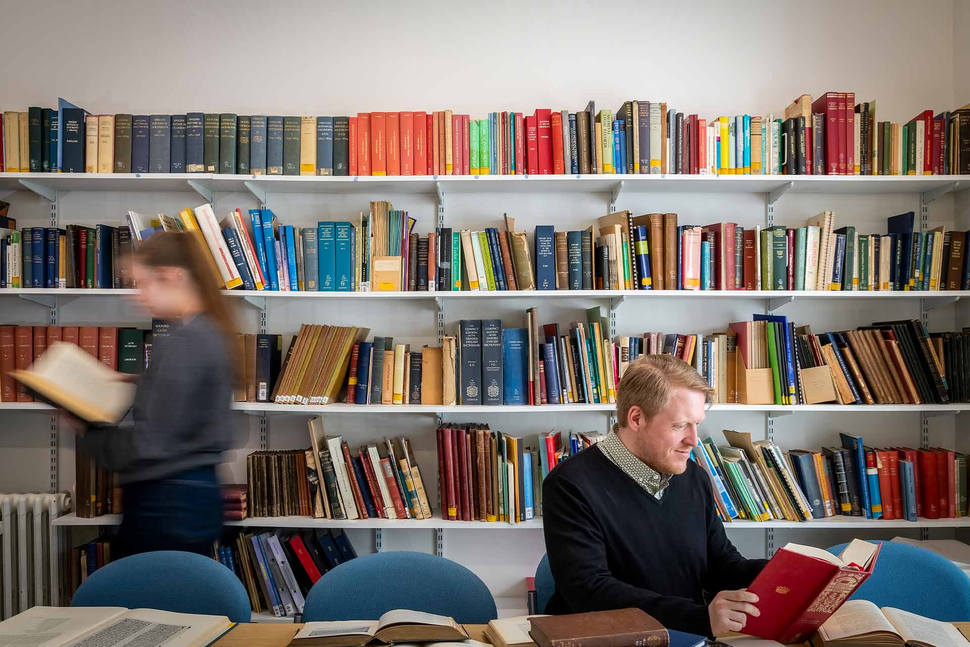 Two people in a library, one walking around while one sat down reading with lots of books piled on the table