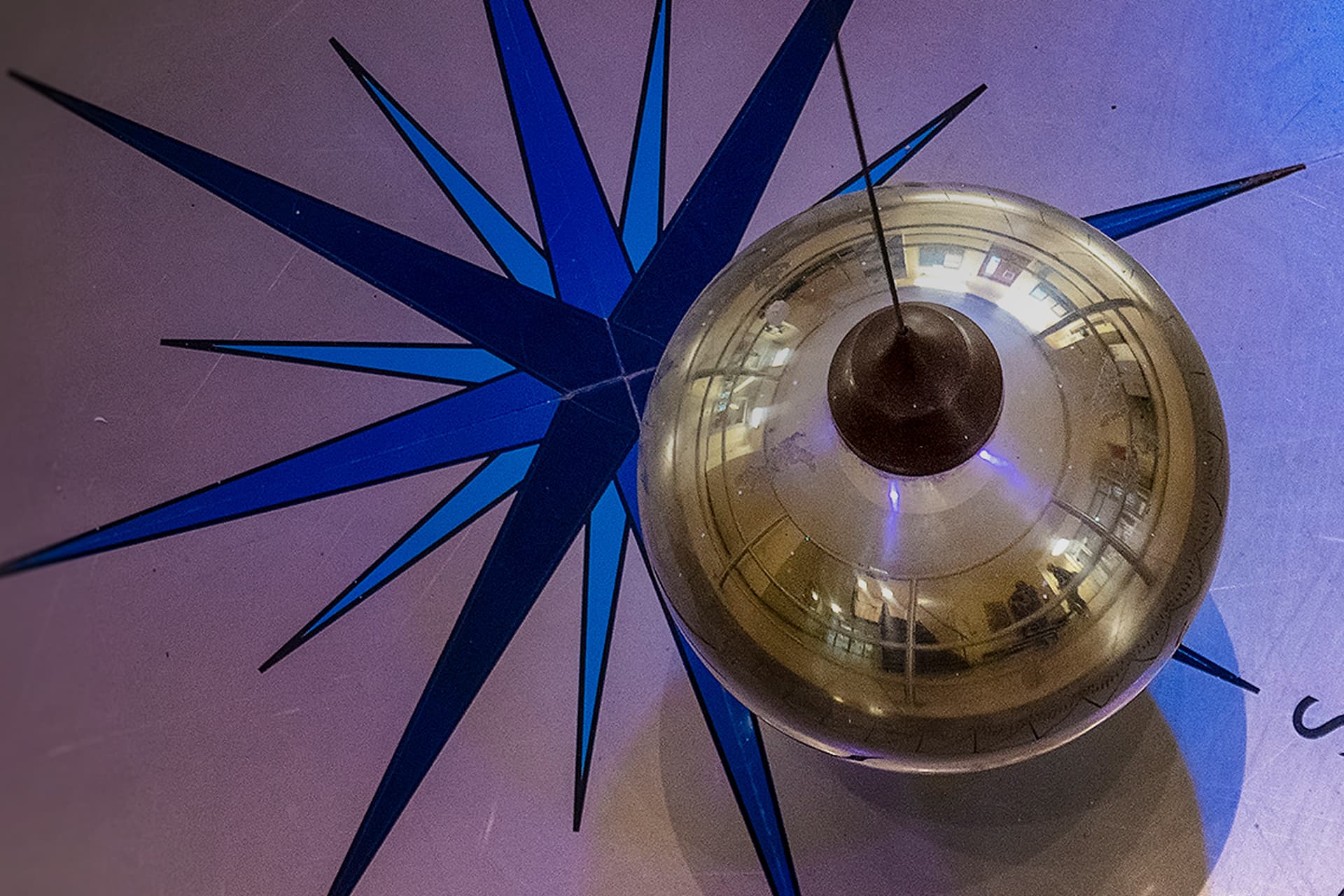 A gold Foucault pendulum over a compass on a tiled floor.