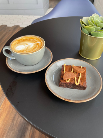 An image of a latte coffee on a table with a square chocolate brownie on a plate.