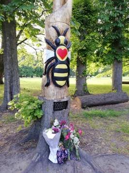 A tree carving of a large bee in a park with bunches of flowers around the base