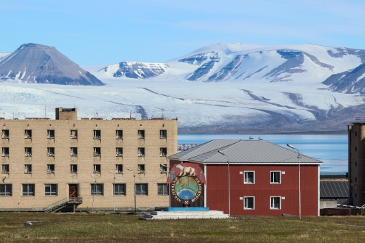 An image of an abandoned red building next to a tall cream building in in front of an arctic vista of a snowy mountain range