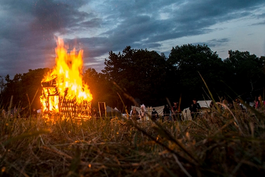 Image of a wooden ram being burnt in a field with people standing watching in front of white tents