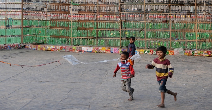 Two small Bangladeshi children chasing a plastic kit in front of a green patterned wall