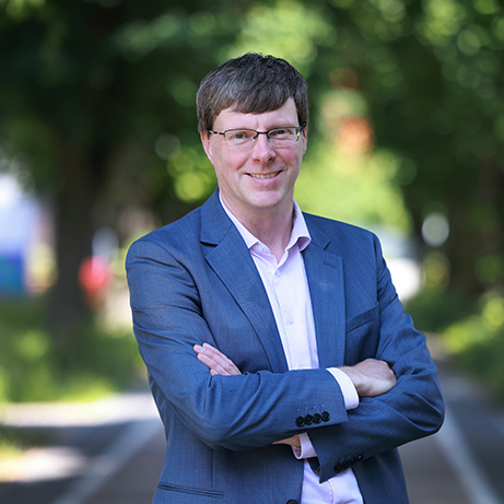 A photograph of Professor Joost Vossebeld smiling in front of a background filled with trees and sunlight