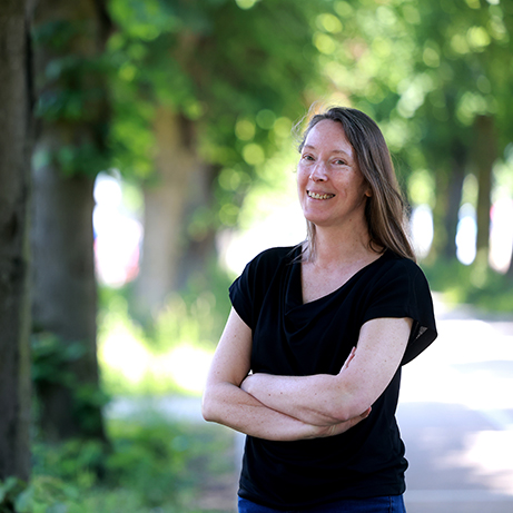 A photograph of Professor Tara Shields smiling in front of a background filled with trees and sunlight