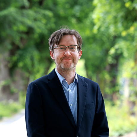 A photograph of Professor Tim Veal smiling in front of a background filled with trees and sunlight