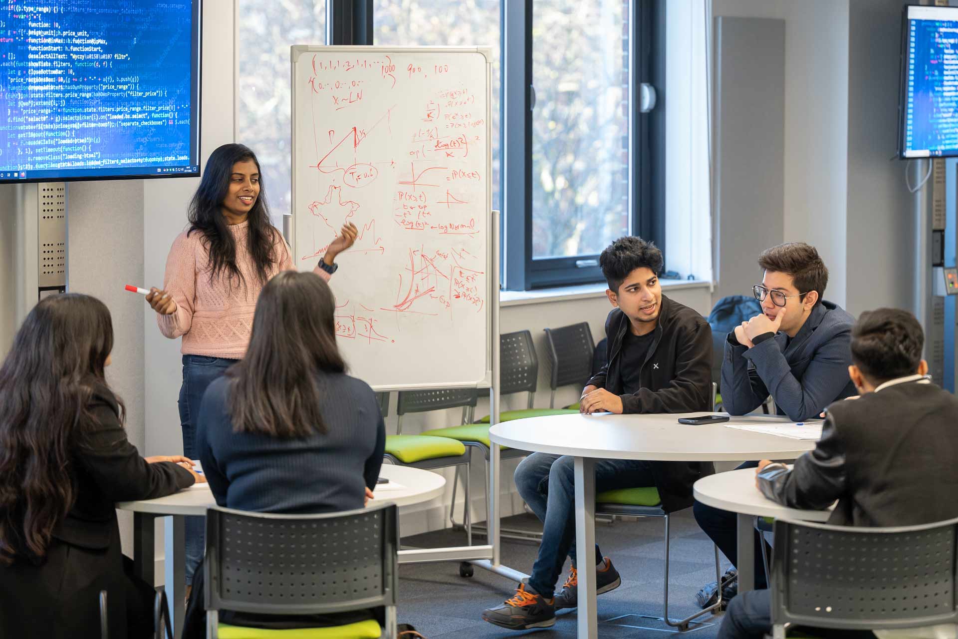 Female student doing a presentation in front of other students with a screen with code and a whiteboard with formulas in the background.